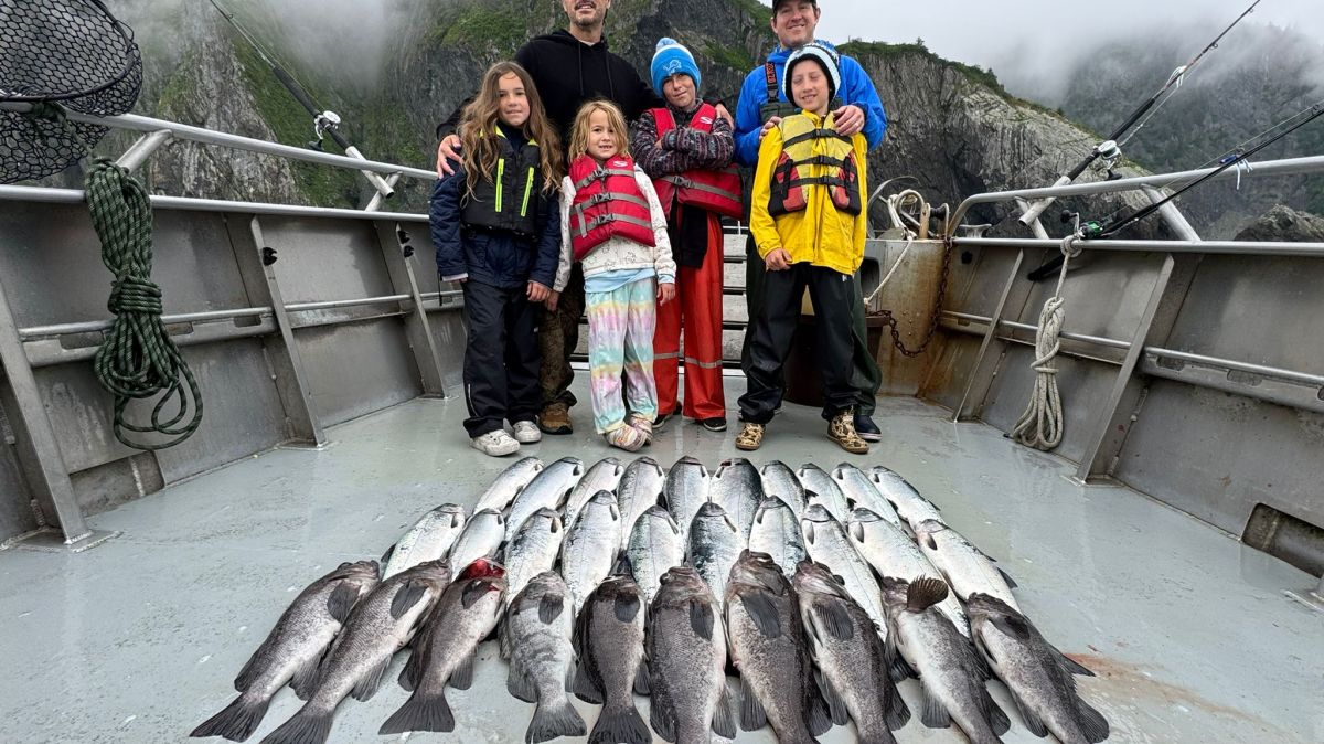 Group on boat with fishing gear and many fish, mountains in background.