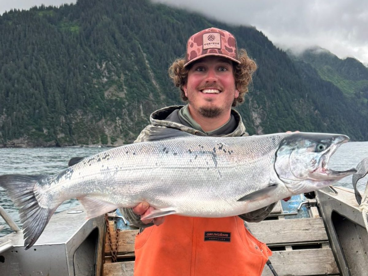 Person in orange overalls holds large fish on a boat with mountains and cloudy sky in background.