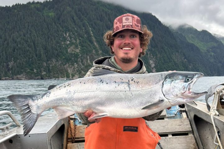 Person in orange overalls holds large fish on a boat with mountains and cloudy sky in background.