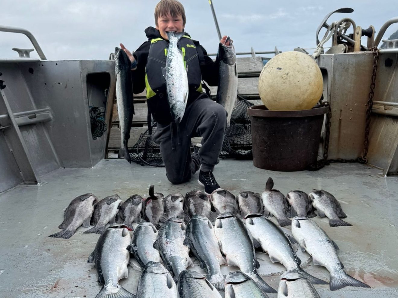 Person on boat holding fish, with many fish lined up on deck, cloudy sky above.