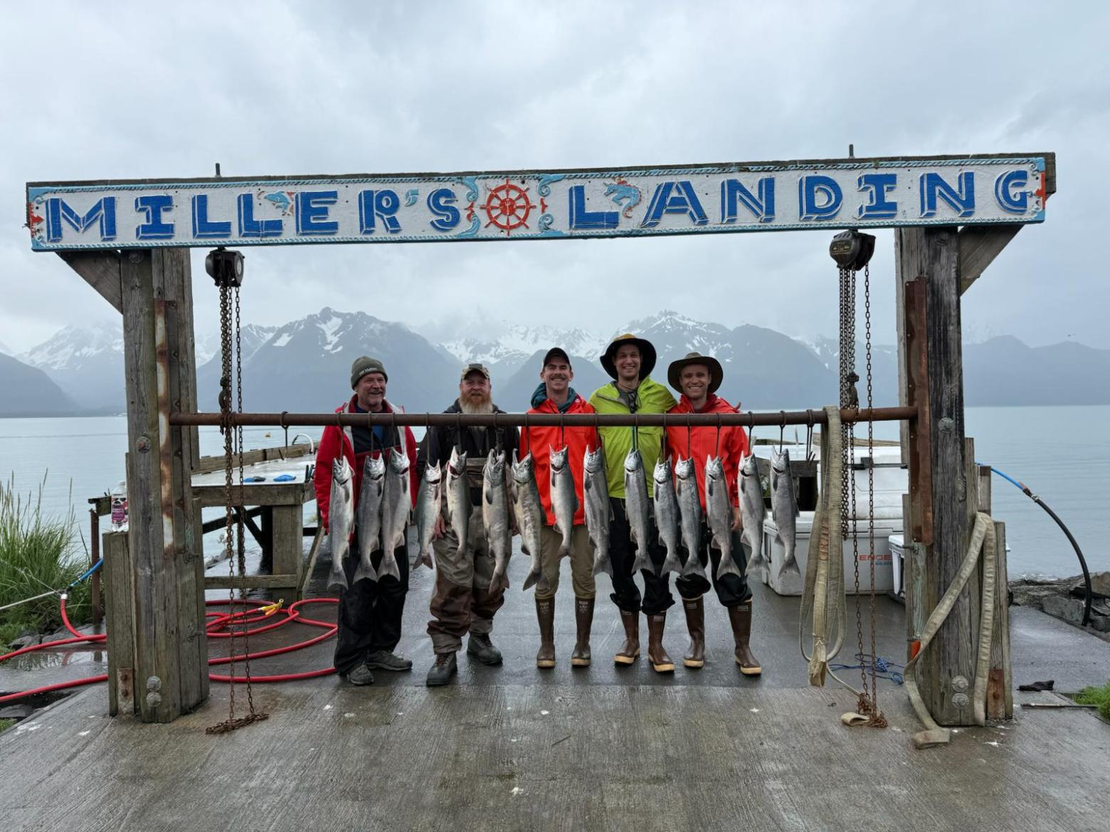 Five people smiling under 'Miller's Landing' sign with caught fish hanging, mountains and water in background.
