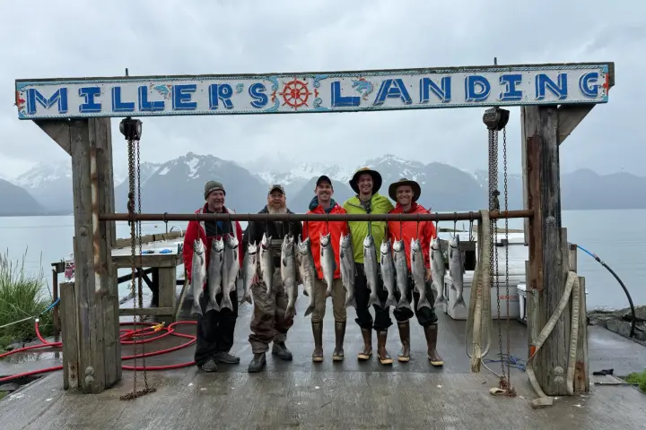 Five people smiling under 'Miller's Landing' sign with caught fish hanging, mountains and water in background.