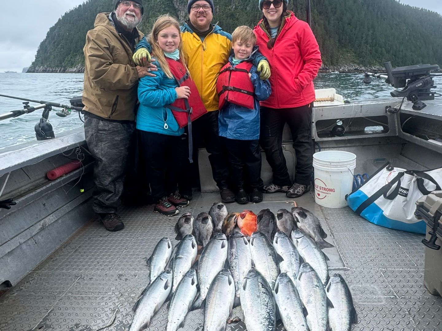 Family on boat with caught fish, smiling with mountain and water backdrop.
