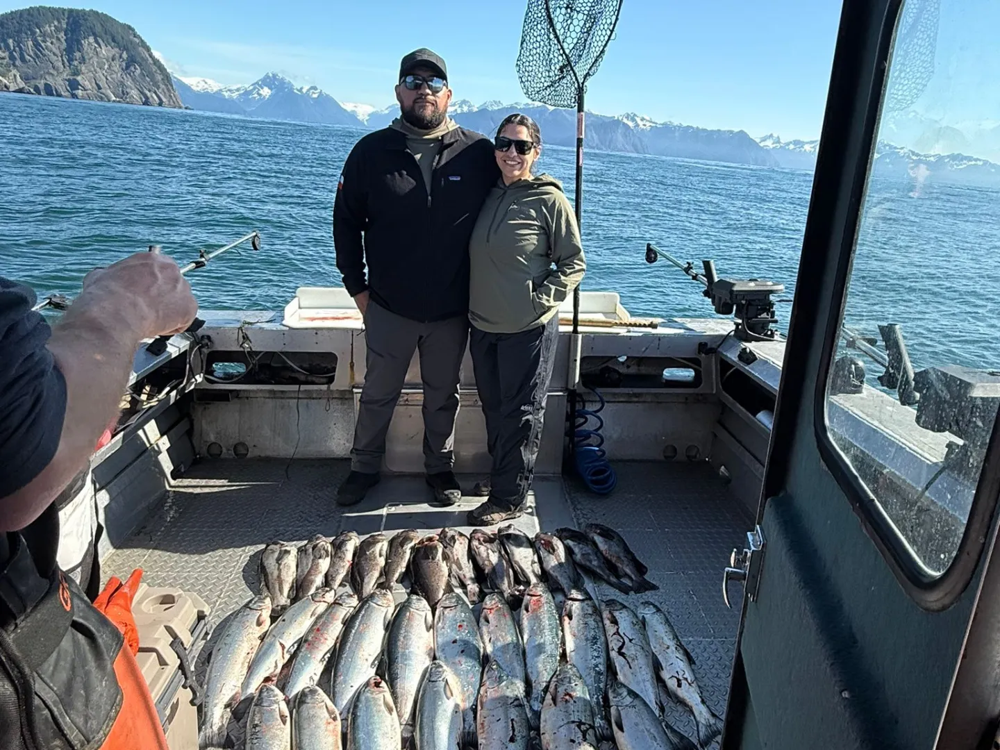 Two people on a boat with a large catch of fish, ocean and mountains in the background.