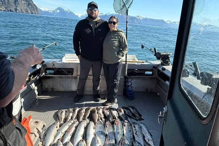 Two people on a boat with a large catch of fish, ocean and mountains in the background.