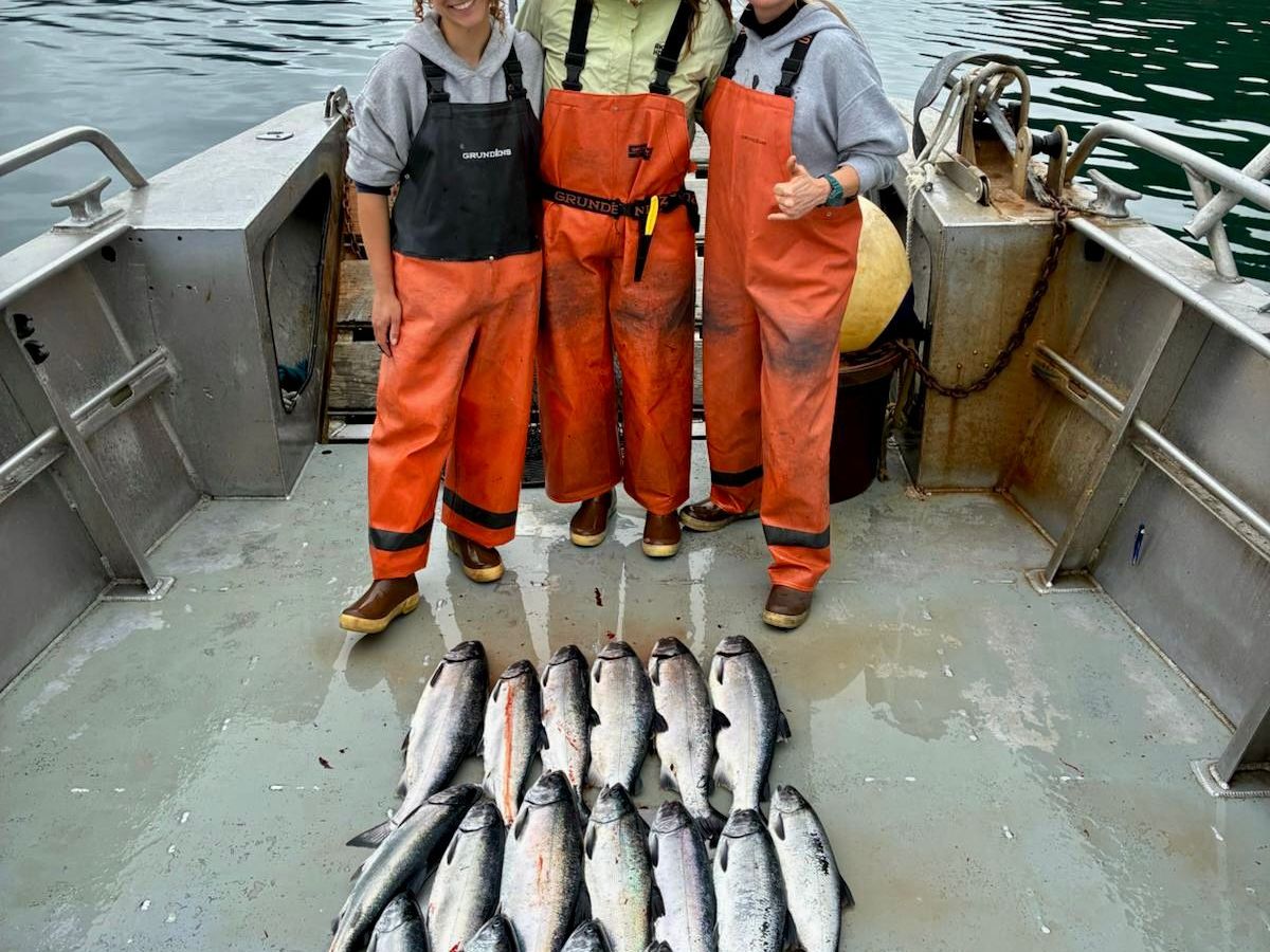 Three people in orange gear with caught fish on a boat, mountains and water in the background.