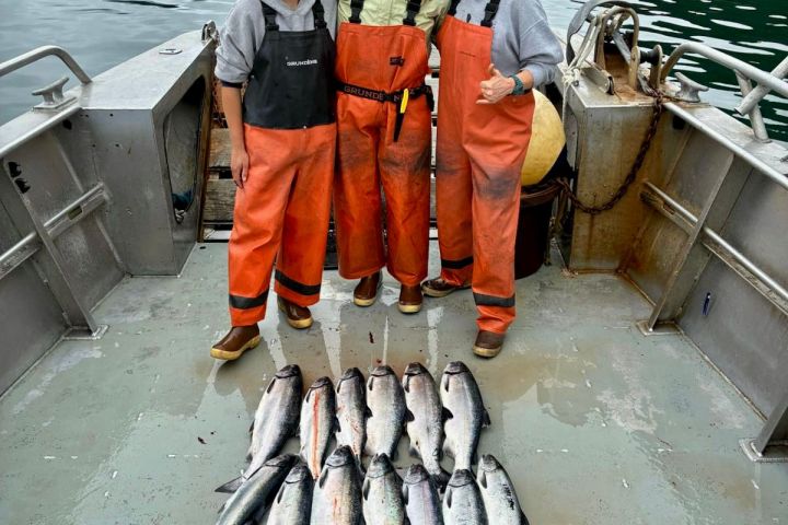 Three people in orange gear with caught fish on a boat, mountains and water in the background.