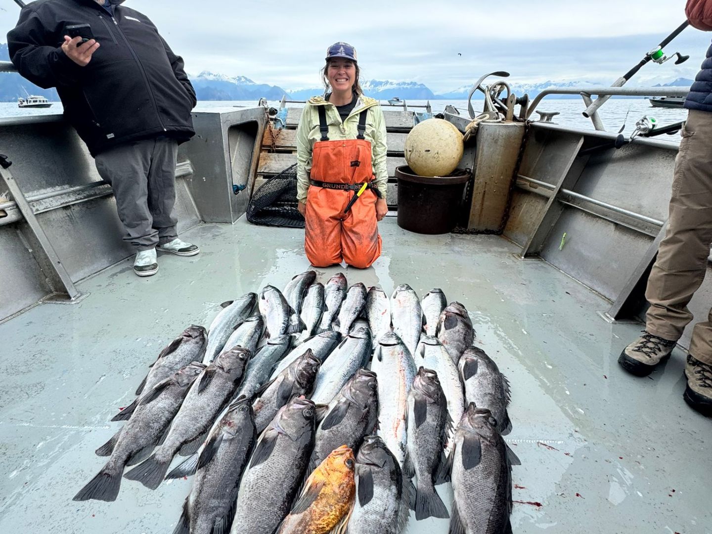 Person kneels on boat deck with a large catch of fish, mountains visible in the background.