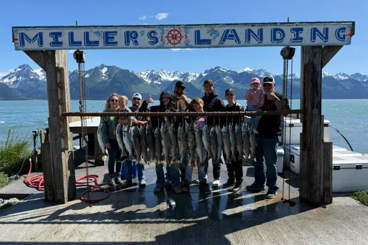 Group of people posing with caught fish under a Miller's Landing sign by a lake with mountains in the background.