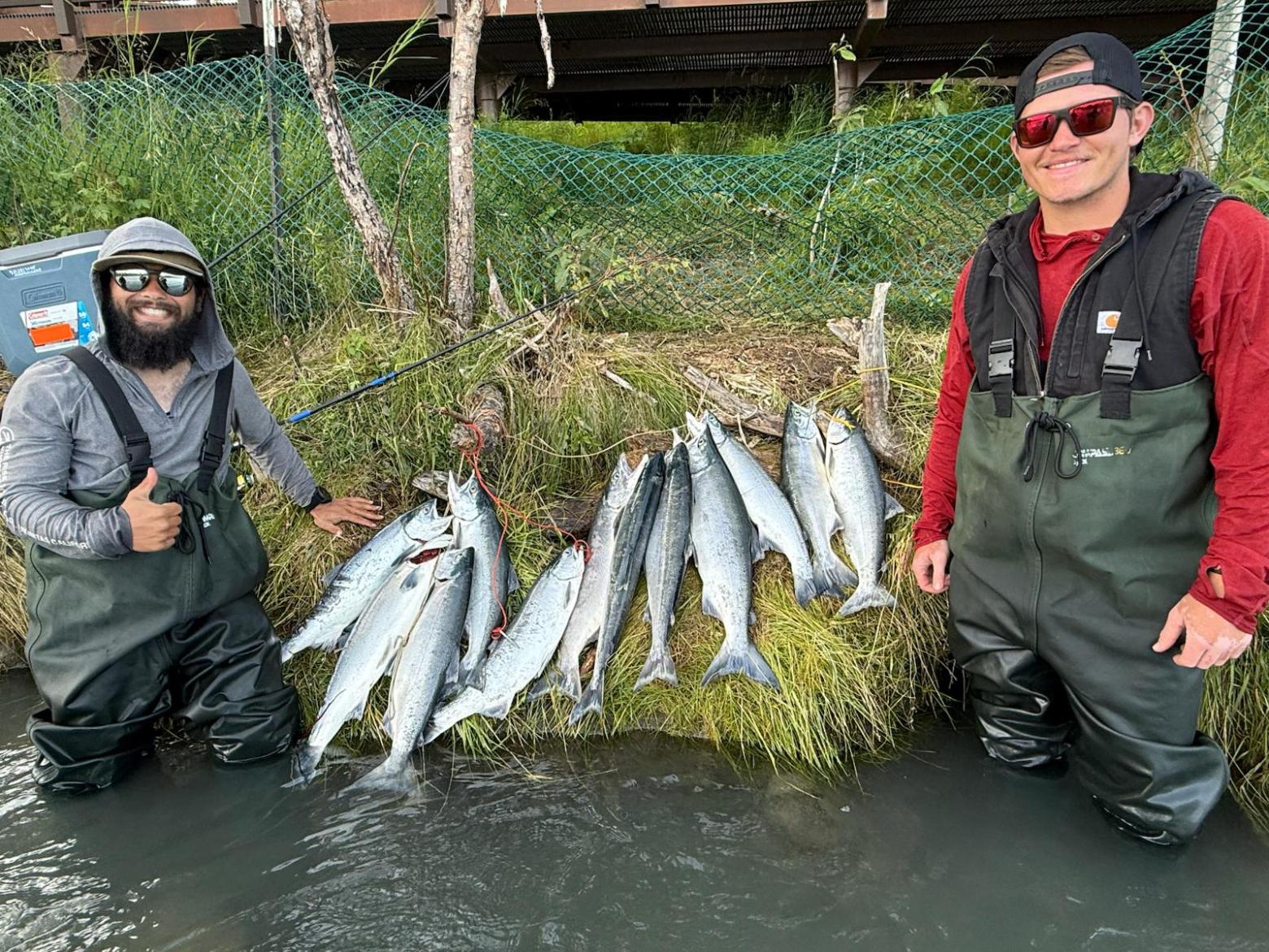 Two people in waders stand by a riverbank with several caught fish displayed on grass.