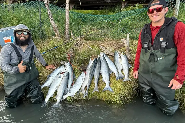 Two people in waders stand by a riverbank with several caught fish displayed on grass.