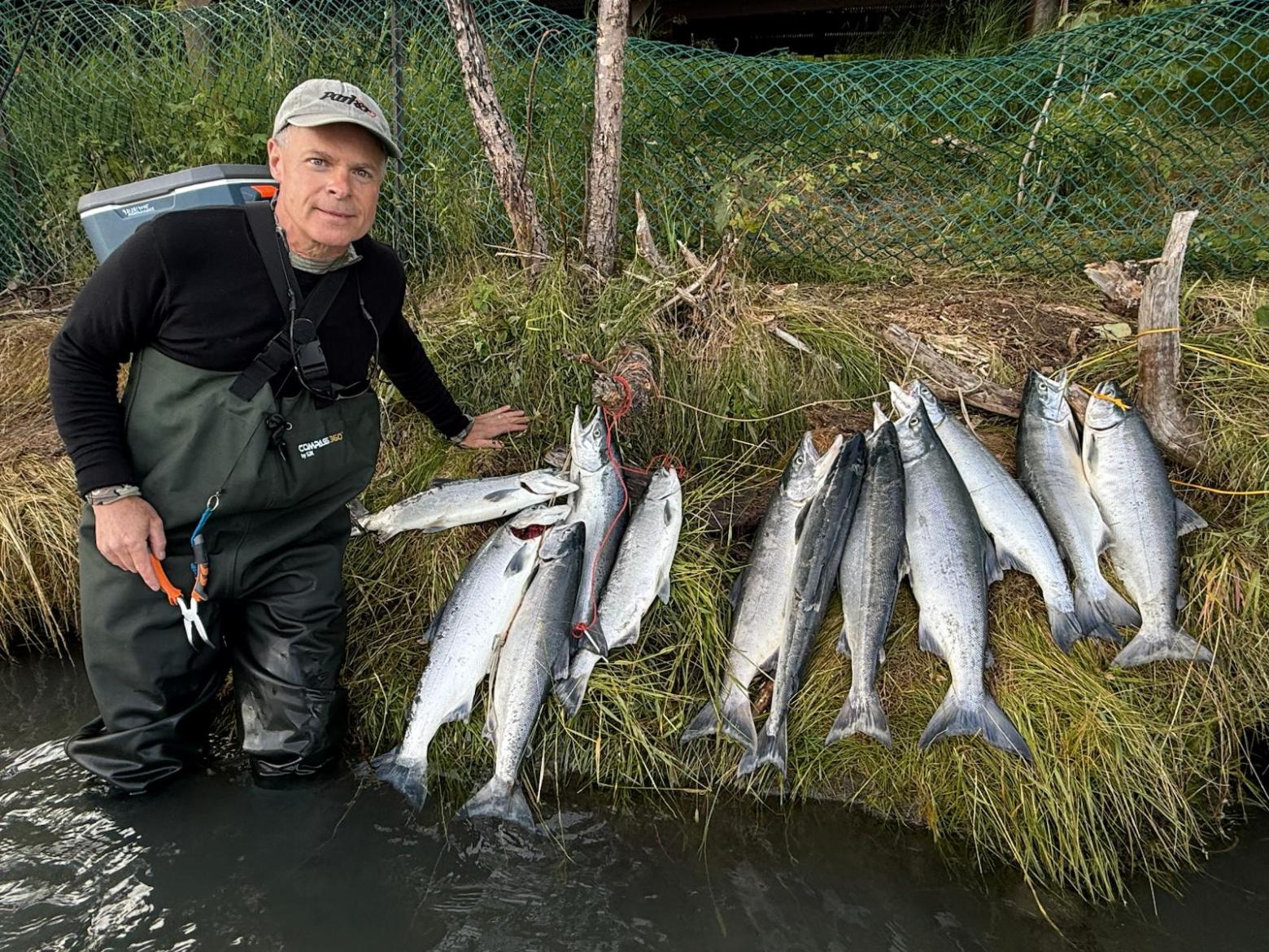 Person stands in water, posing with a catch of eight fish on the grass beside them.