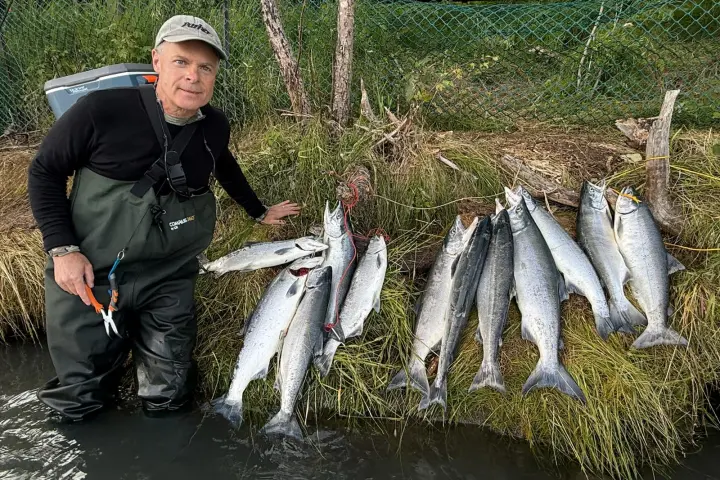 Person stands in water, posing with a catch of eight fish on the grass beside them.