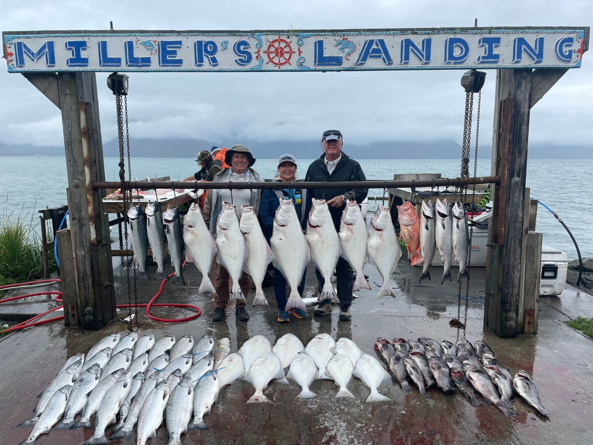 Three people stand by a sign labeled 'Miller's Landing' with numerous large fish hanging and lying beneath.