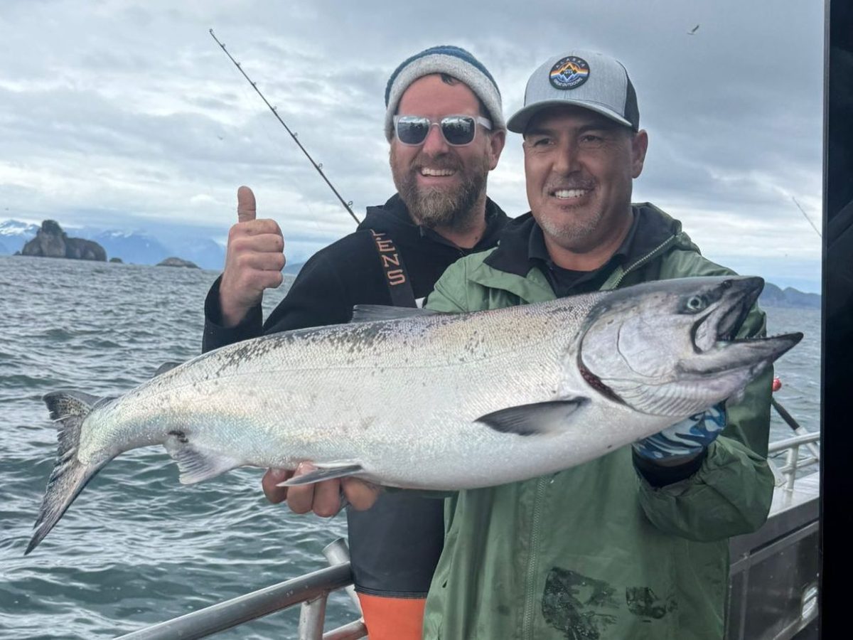 Two men on a boat holding a large fish, with one giving a thumbs up. Overcast sky and mountains in the background.