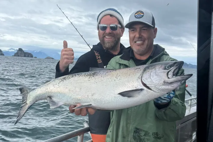 Two men on a boat holding a large fish, with one giving a thumbs up. Overcast sky and mountains in the background.