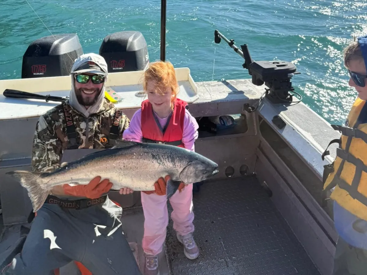 Child and adult holding a large fish on a boat with mountains and lake in the background.