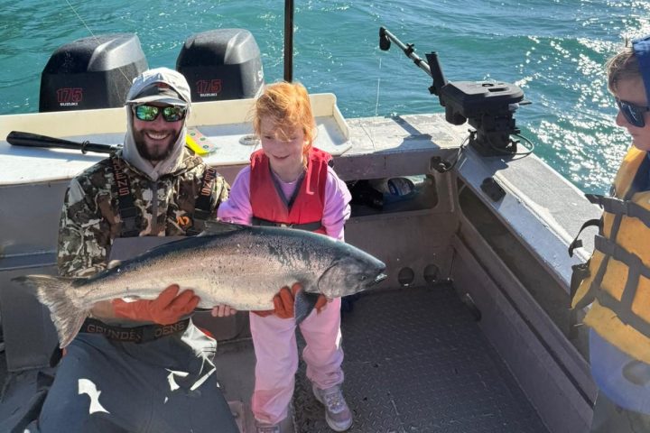 Child and adult holding a large fish on a boat with mountains and lake in the background.