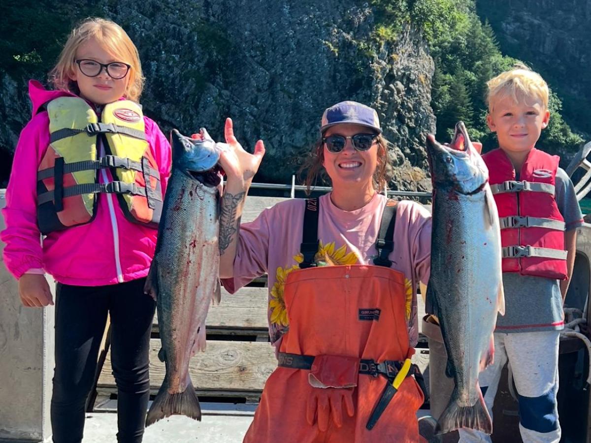 Person kneeling holds up two fish, flanked by two children in life vests on a boat.
