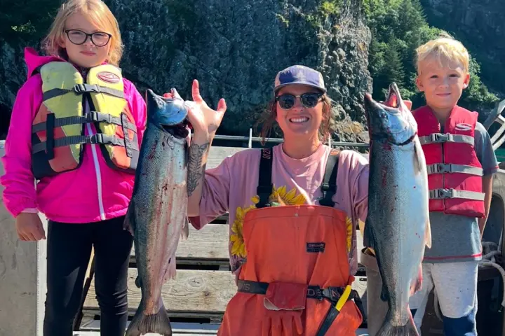 Person kneeling holds up two fish, flanked by two children in life vests on a boat.