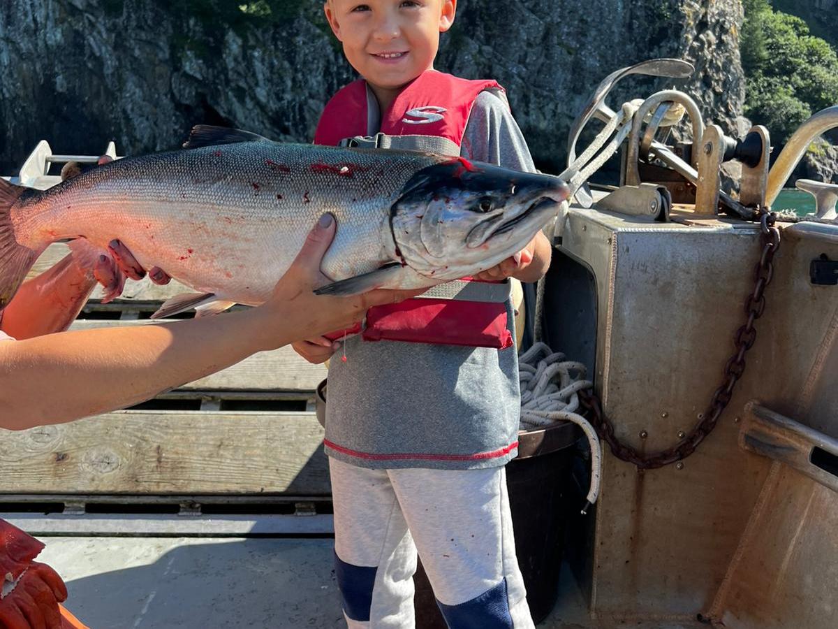 Child in red life jacket holding a large fish on a boat with rocky cliffs in the background.