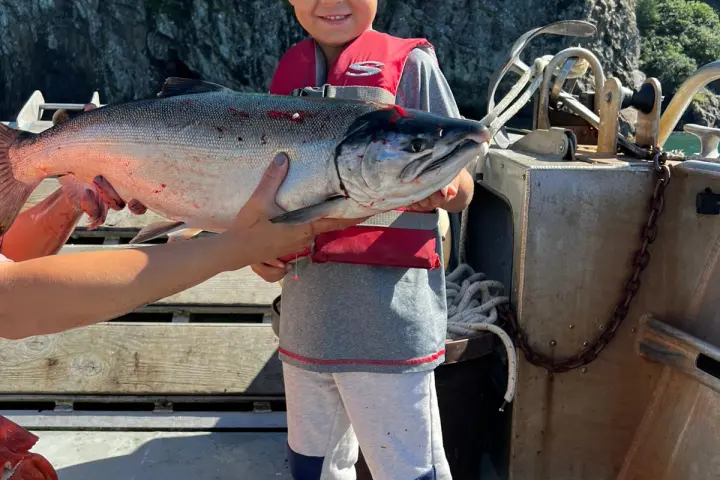 Child in red life jacket holding a large fish on a boat with rocky cliffs in the background.