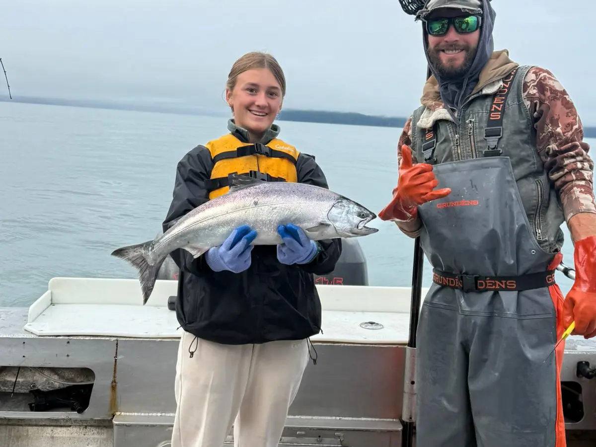 Two people on a boat holding a large fish, with one wearing gloves and the other in a fishing net.