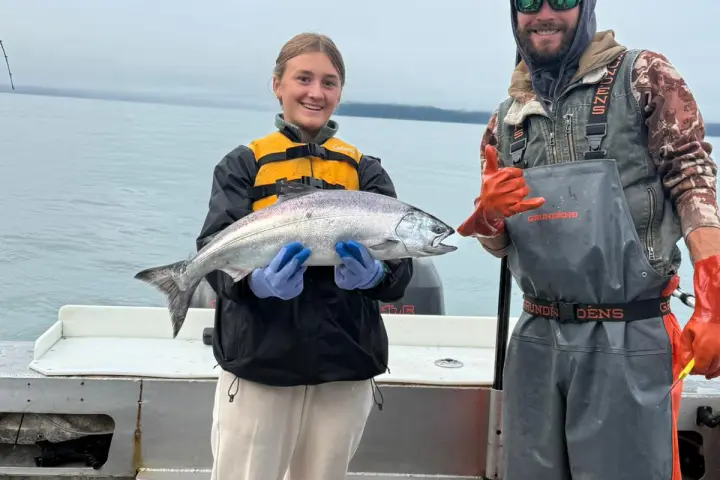 Two people on a boat holding a large fish, with one wearing gloves and the other in a fishing net.