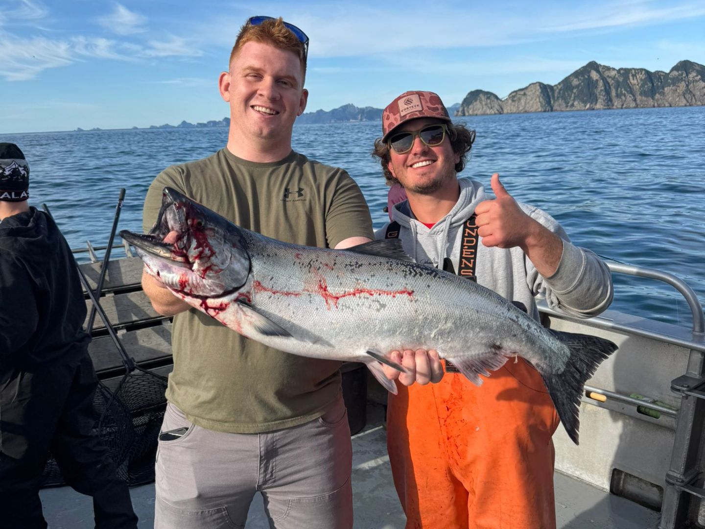 Two men on a boat holding a large fish, with ocean and mountains in the background.