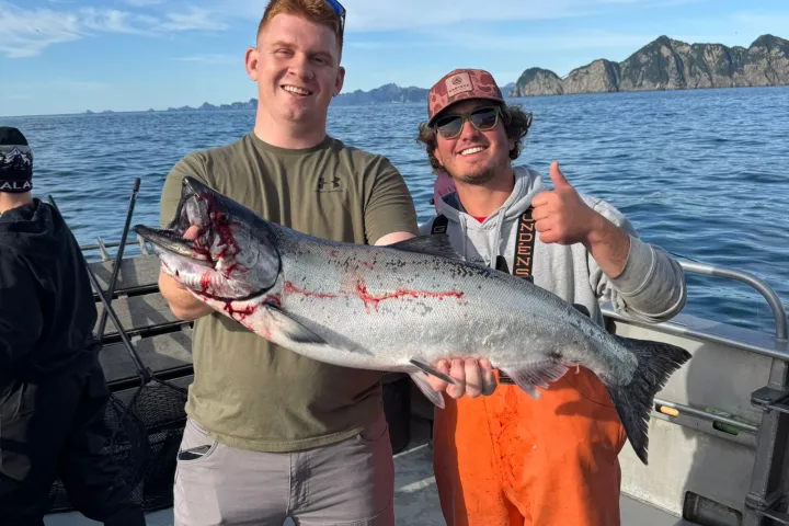 Two men on a boat holding a large fish, with ocean and mountains in the background.
