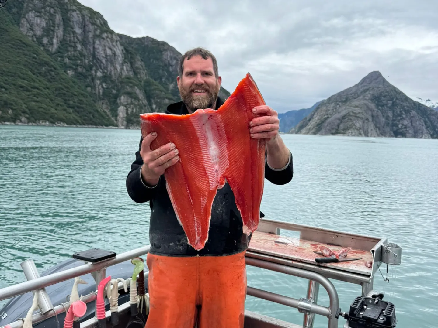Person holding large fish fillet on a boat with mountains and water in the background.