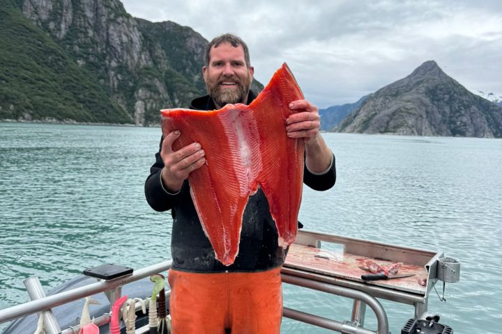 Person holding large fish fillet on a boat with mountains and water in the background.
