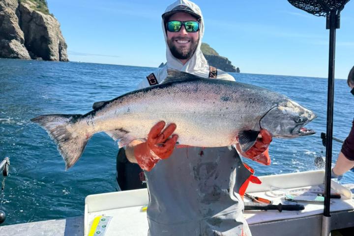 Person holding large fish on boat with ocean and cliffs in the background.