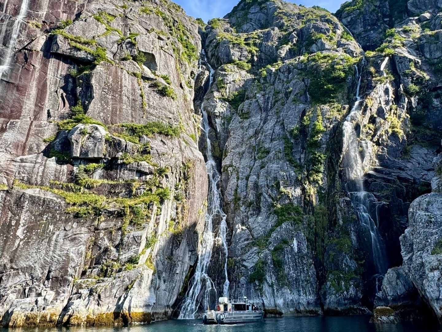 Boat on turquoise water with towering rocky cliffs and small waterfalls under a blue sky.