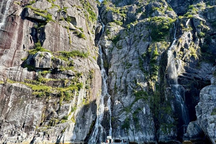 Boat on turquoise water with towering rocky cliffs and small waterfalls under a blue sky.