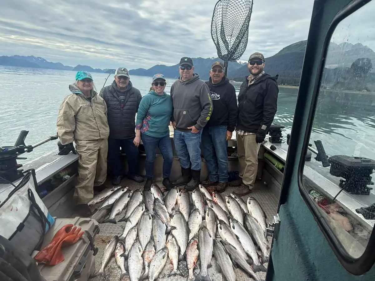 Six people on a boat with a large catch of fish on the deck, mountains and water in the background.