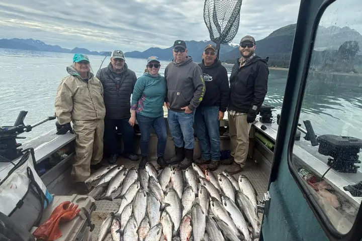 Six people on a boat with a large catch of fish on the deck, mountains and water in the background.