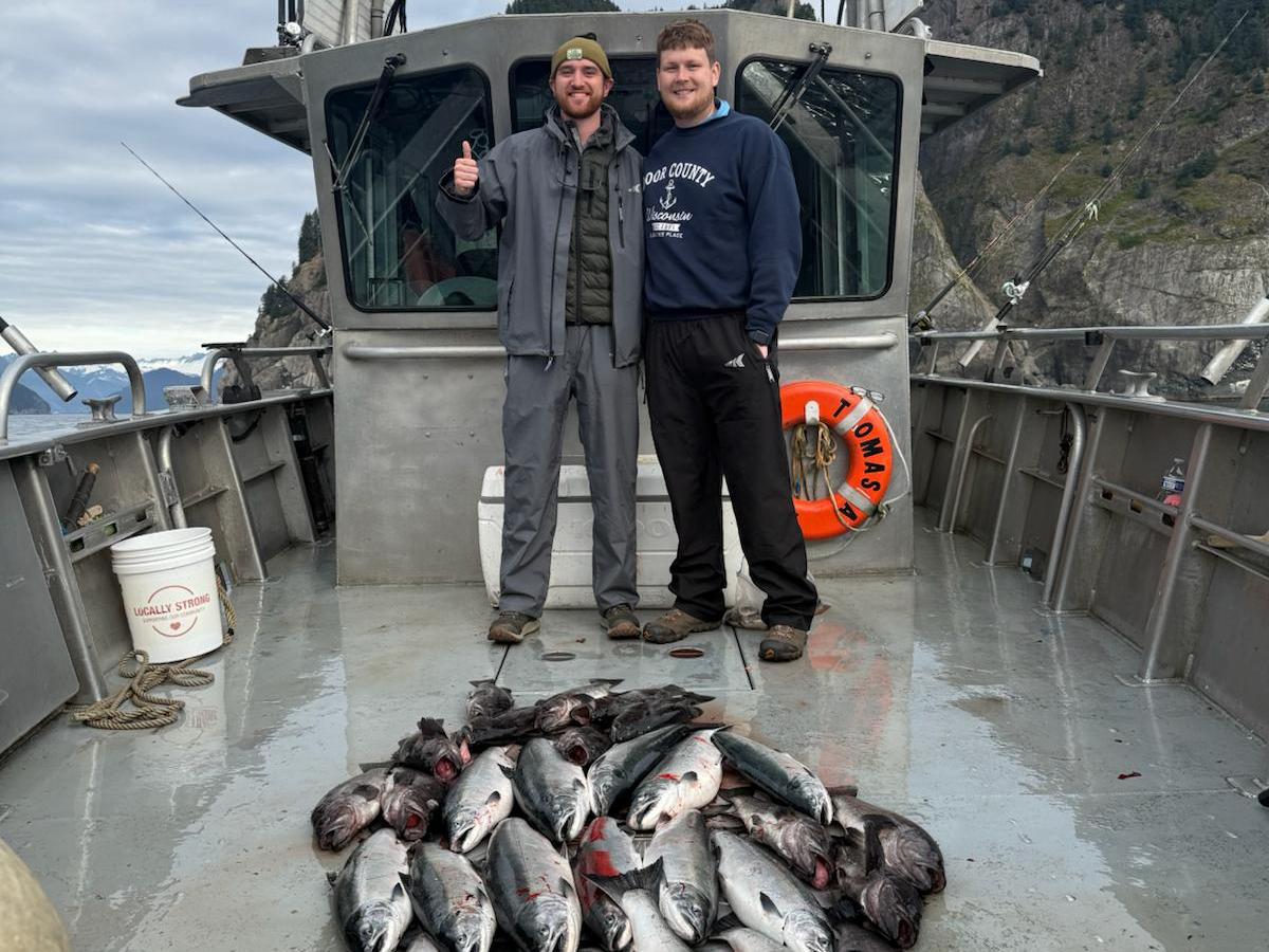 Two people on a boat with a large catch of fish, mountains in the background.
