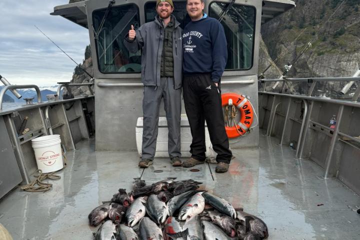 Two people on a boat with a large catch of fish, mountains in the background.