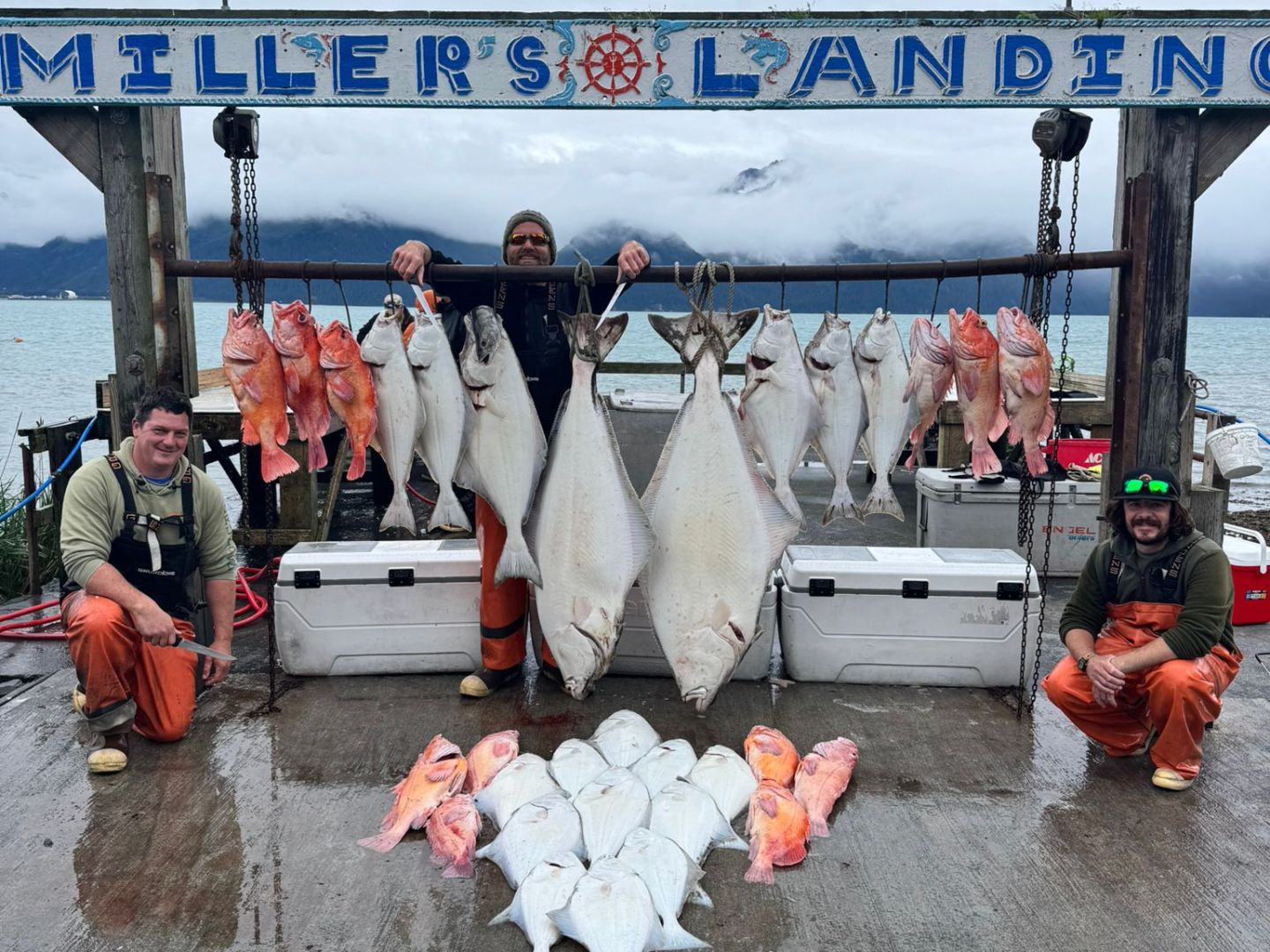 Three people display large fish catch at Miller's Landing with mountains and water in the background.