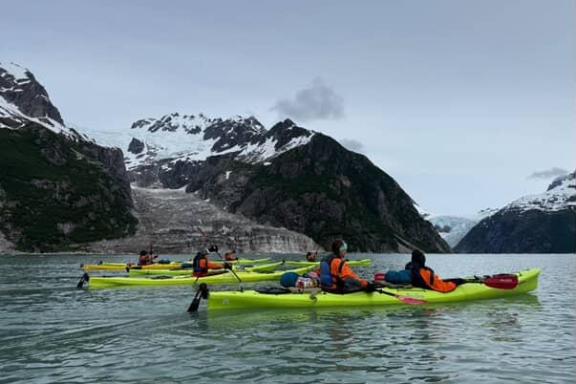 People kayaking on a lake near snow-capped mountains under a cloudy sky.