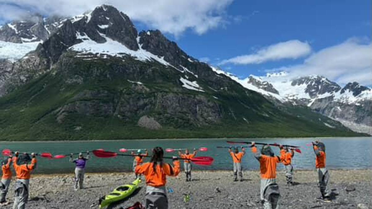 Group in orange jackets holding paddles by a lake with snow-capped mountains in background.
