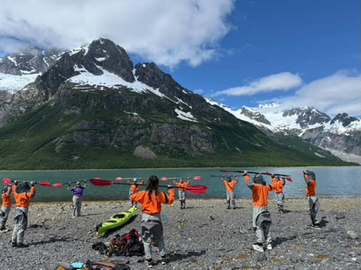 Group in orange jackets holding paddles by a lake with snow-capped mountains in background.