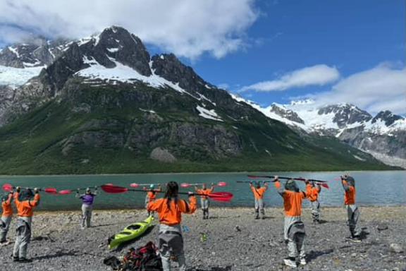 Group in orange jackets holding paddles by a lake with snow-capped mountains in background.
