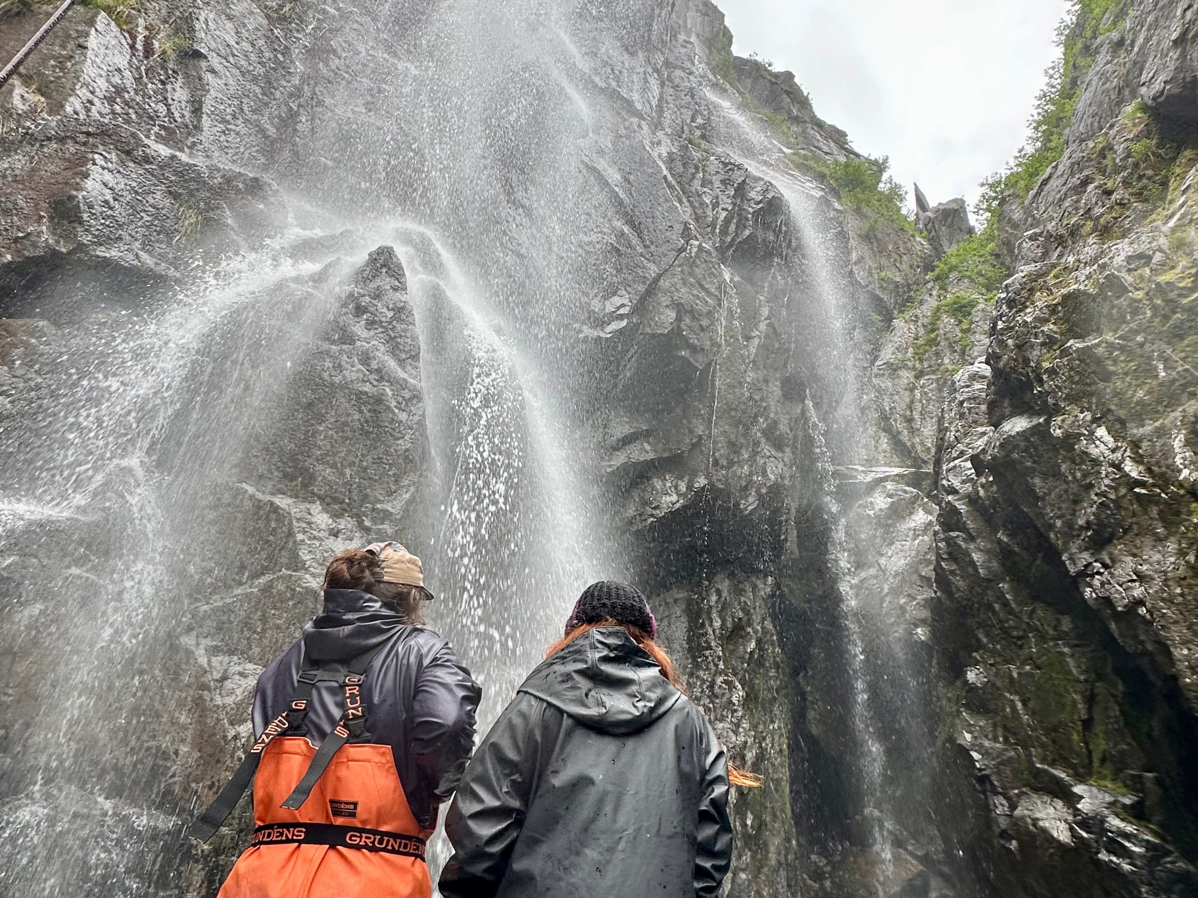 Two people in raincoats looking up at a tall waterfall on a rocky cliff.