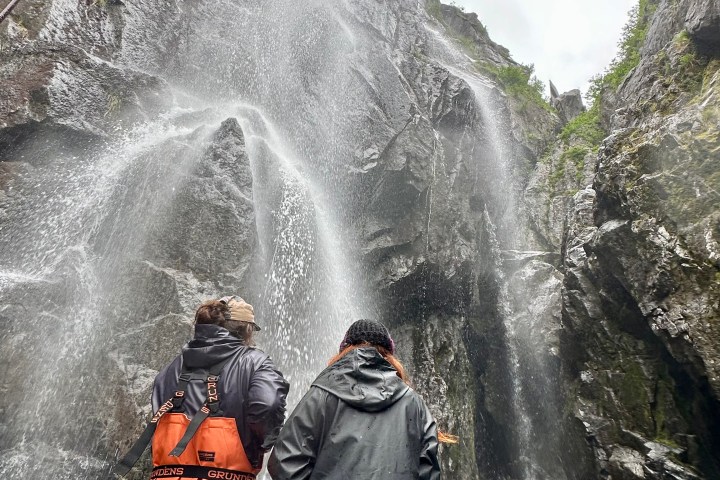 Two people in raincoats looking up at a tall waterfall on a rocky cliff.