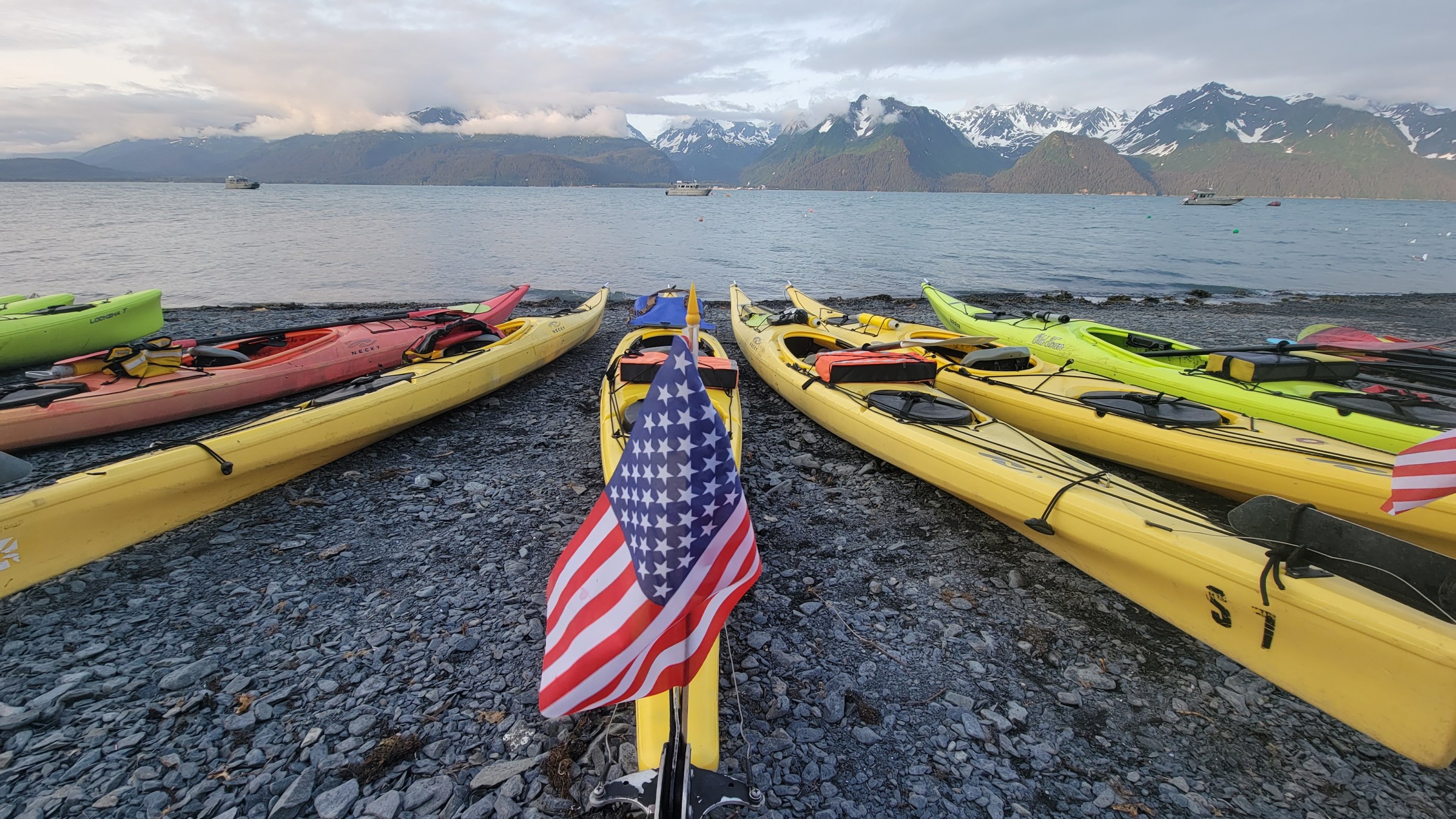 Colorful kayaks with American flags on rocky shore, mountainous backdrop and calm sea.
