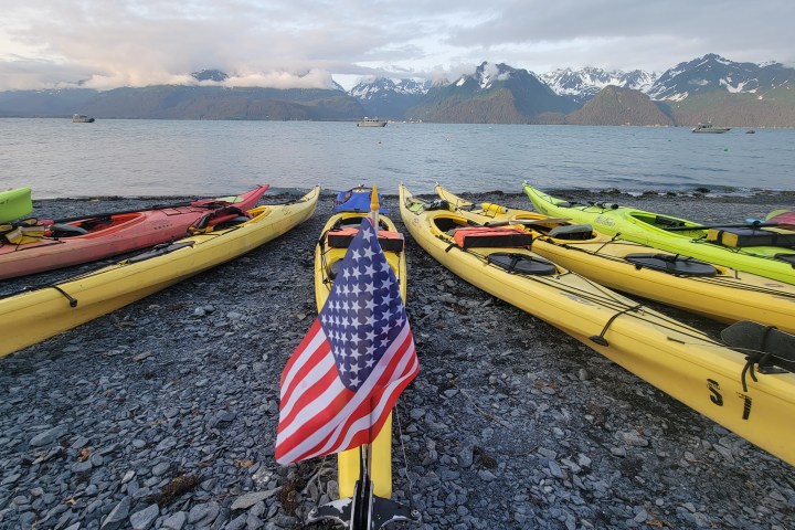 Colorful kayaks with American flags on rocky shore, mountainous backdrop and calm sea.