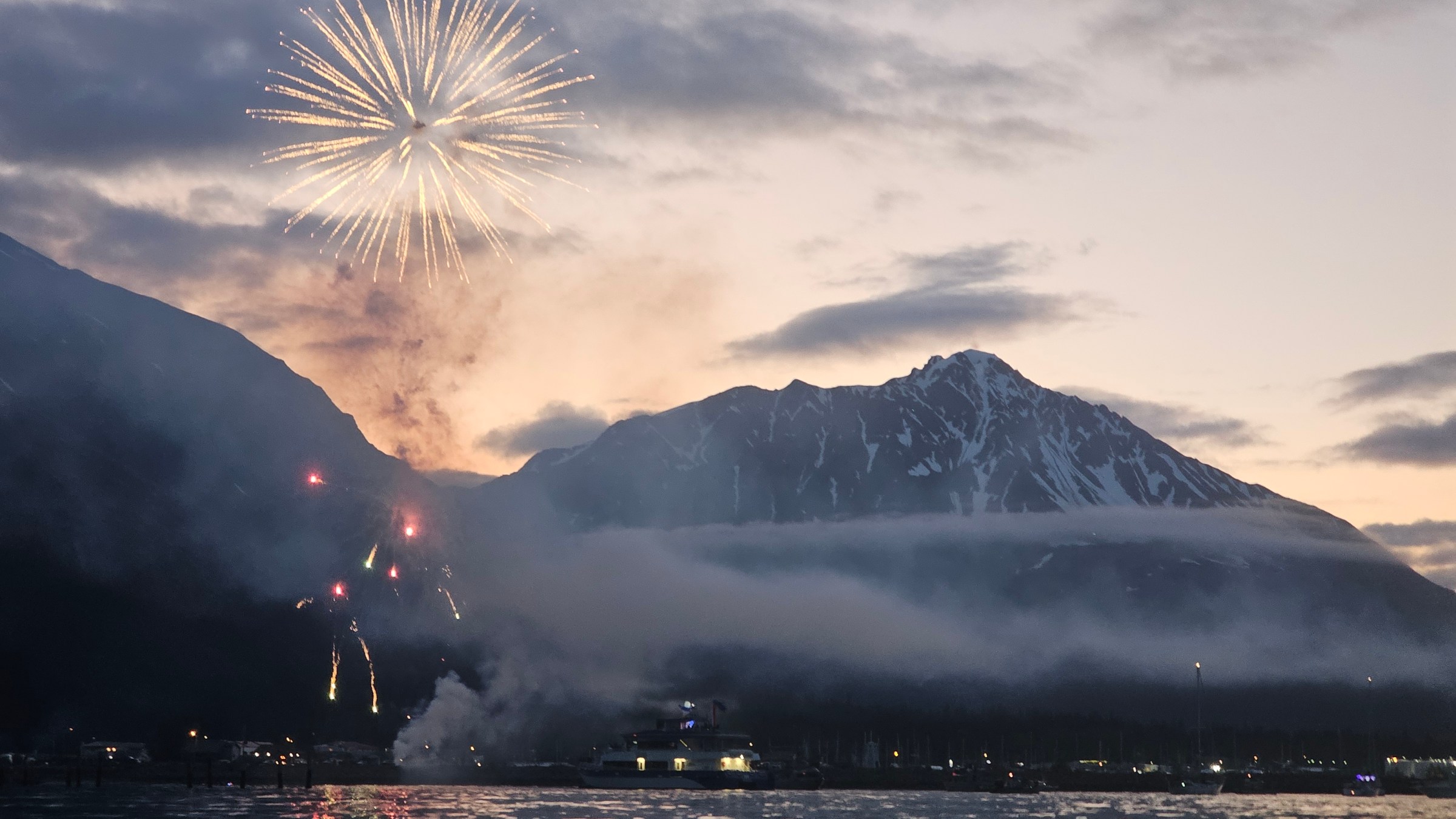 Fireworks lighting up a mountainous landscape at dusk, with clouds and a body of water in the foreground.