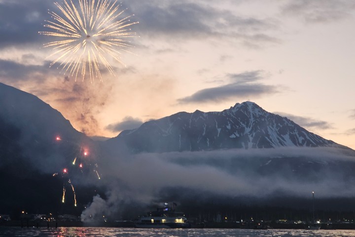 Fireworks lighting up a mountainous landscape at dusk, with clouds and a body of water in the foreground.
